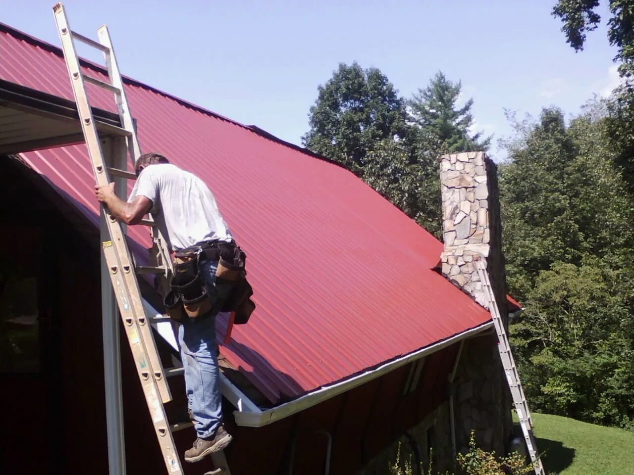 Roofer on a ladder beside a red metal roof and stone chimney — Soffit & Fascia Repair by Roofing Brothers in Newark Valley